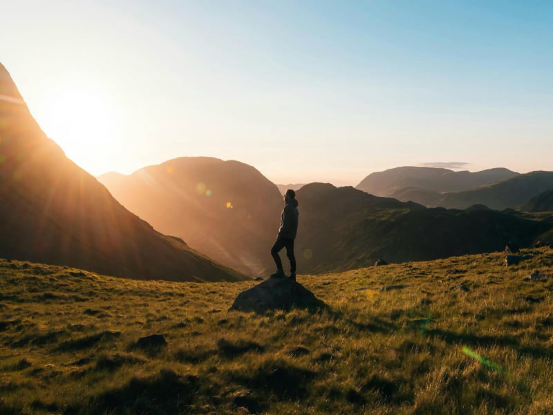 The Best UK Destinations for walkers. A sunset over hills with a walker stood on a rock. The Outdoor Guide.