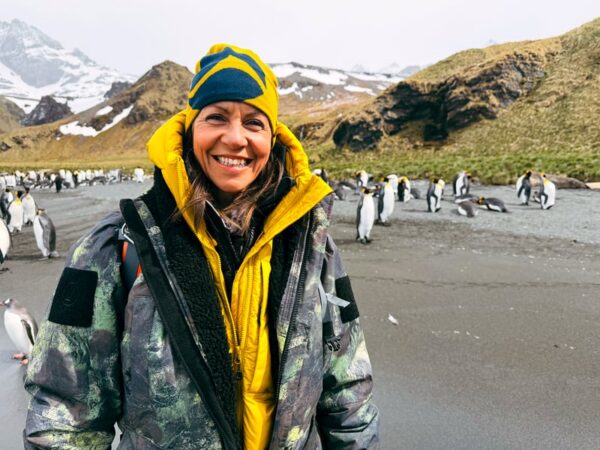 Wonders of the Frozen South ITV Episode 2 - South Georgia. Julia Bradbury stands with penguins, she iw earing a ThruDark jacket and bright yellow jacket.
