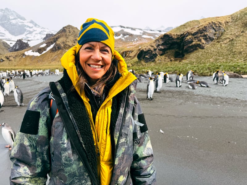 Wonders of the Frozen South ITV Episode 2 - South Georgia. Julia Bradbury stands with penguins, she iw earing a ThruDark jacket and bright yellow jacket.